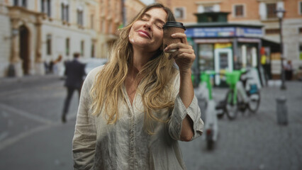 Fototapeta premium Woman holding coffee cup to face, eyes closed and smiling on a cobblestone street; contentment morning ritual.
