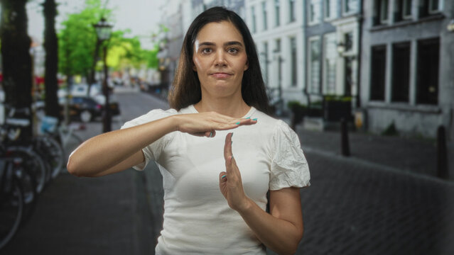 Young hispanic woman in a casual white top with hands making timeout gesture on a sunny urban street lined with bicycles; determination.