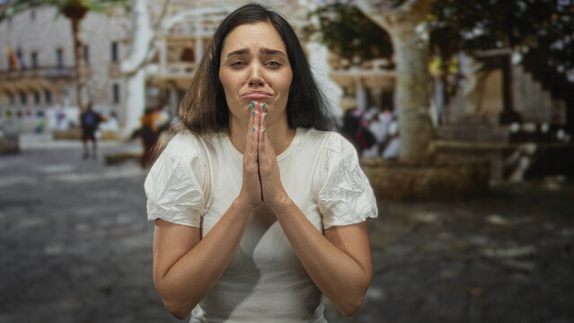 Woman with hands pressed in prayer gesture on bustling public cobblestone street beside stone building; desperation.