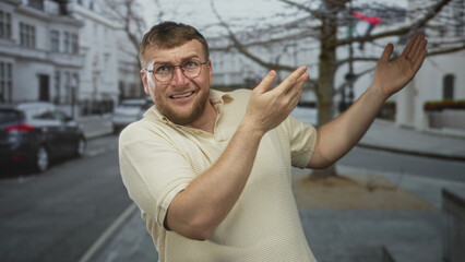 Fototapeta premium Man wearing glasses and a beige polo gesturing with both hands and open palms on a city street near parked cars and white buildings; surprise awkwardness.