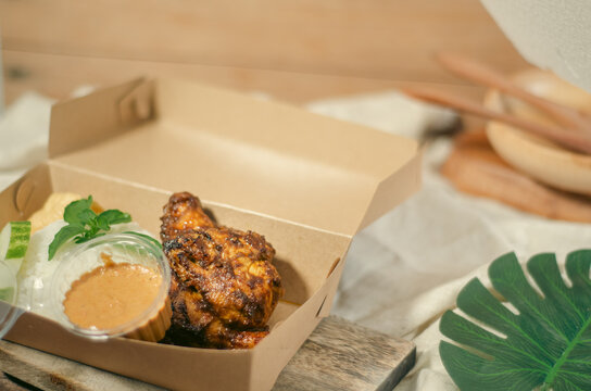 Indonesian ayam bakar bumbu rujak served with rice and sambal in eco takeaway box. Rustic food styling with warm tones, traditional cuisine concept.