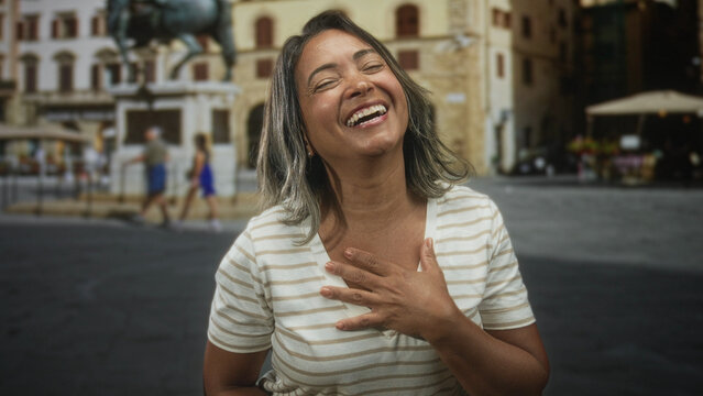 Hispanic woman with hand on chest laughing in front of a historic building plaza, striped shirt and earring visible; joy gratitude.