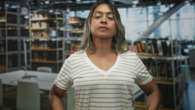 Woman middle age wearing glasses with hands on hips and bare shoulders adjusting striped shirt in a library building; calm confidence.