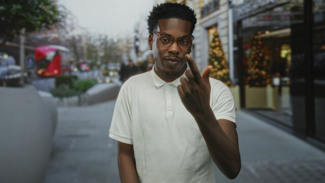 Man in white polo shirt beckons with finger on urban city street near building facade; friendly invitation.