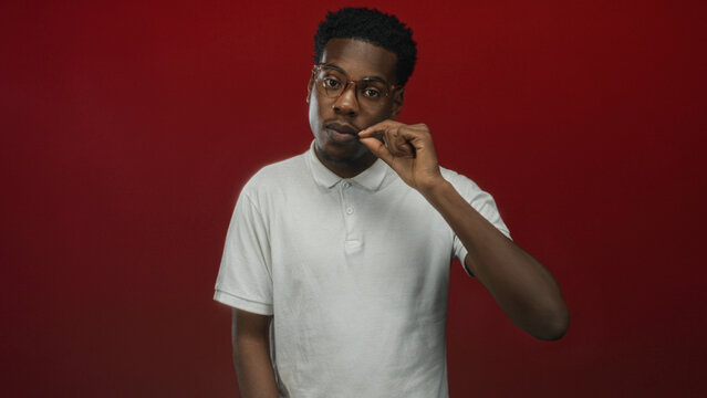 Young african american man wearing glasses and white polo with hand covering mouth for silence in red studio setting; silence.