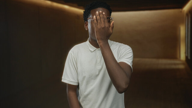 Man covers face with hand in studio corridor with warm lighting and textured walls while wearing polo shirt and glasses, scrunched expression; mystery.