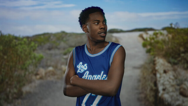 Man wearing blue los angeles jersey with crossed arms on quiet dirt street under cloudy sky; rejection.