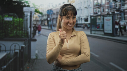 Fototapeta premium Woman with crossed arms and slight smirk wearing beige zip top on a city street near a bus shelter and railing; confidence assertiveness.