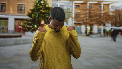 Naklejka premium Young black man in yellow sweater raises his fists in a decorated street with a large christmas tree; quiet triumph.
