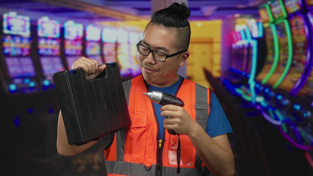 Man holding black toolbox and electric screwdriver in casino building while smiling and posing, wearing reflective vest; skilled maintenance confidence.