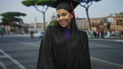 Naklejka premium Young latina woman in black graduation cap and gown on city street sticks out tongue with playful head tilt; achievement pride.