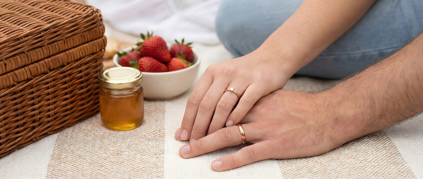 Close up of married couple holding hands with wedding rings on striped blanket, romantic summer picnic with wicker basket, fresh strawberries and honey jar, panoramic banner