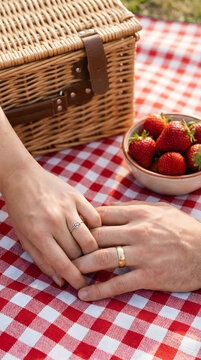 Close up of married couple holding hands with wedding rings on red checkered blanket, romantic summer picnic with wicker basket and fresh strawberries