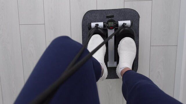 Top view of Woman's Feet in White Sneakers Using Mini Stepper Exercise Machine with Resistance Bands on Gray Mat at Home, Female Legs in Blue Leggings Working Out on Compact Stair Stepper Indoors