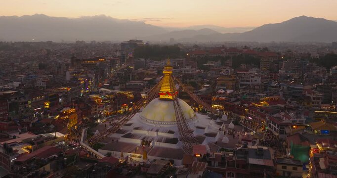 Aerial view of Boudhanath Stupa, UNESCO World Heritage Site in Kathmandu Nepal, glowing at twilight with prayer flags, surrounding crowd and dense city buildings under golden evening light.