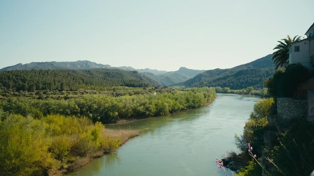 The Ebro River curves past Miravet, Spain, between green riverbanks, forested hills, and distant mountains, creating a wide and peaceful Mediterranean landscape.