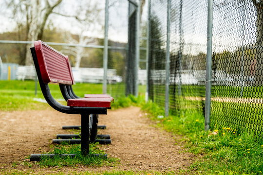 Two empty red metal benches sit along the edge of a baseball field with a chain link fence and green field visible behind them, waiting for players or spectators.