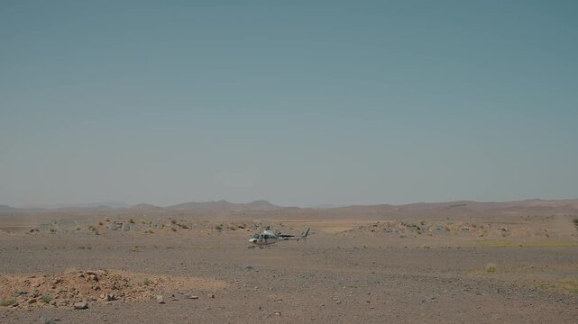 Small white helicopter parked on flat arid desert ground with rocky mountains and hazy sky in background