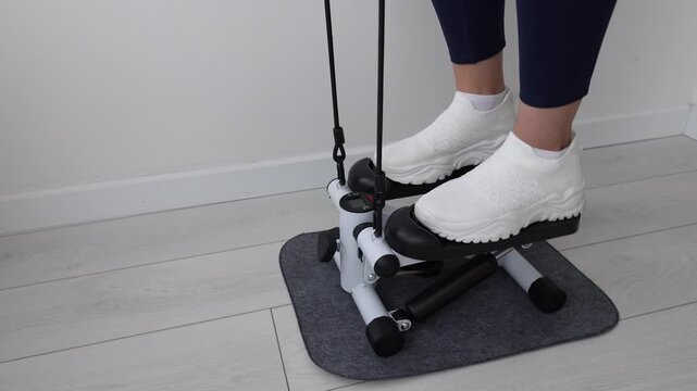 Close-Up of Woman's Feet in White Sneakers Using Mini Stepper Exercise Machine with Resistance Bands on Gray Mat at Home, Female Legs in Blue Leggings Working Out on Compact Stair Stepper Indoors