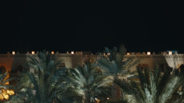 Illuminated clay kasbah wall with decorative battlements and lanterns rising above date palm trees at night