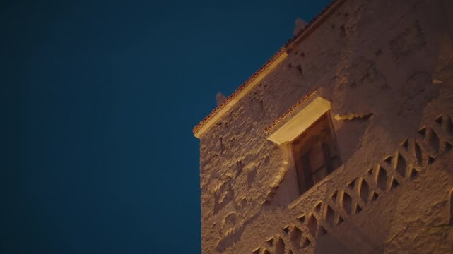 Low angle view of lit clay kasbah tower with carved geometric decoration and wooden window ledge against deep blue night sky