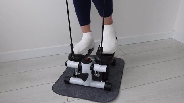 Close-Up of Woman's Feet in White Sneakers Using Mini Stepper Exercise Machine with Resistance Bands on Gray Mat at Home, Female Legs in Blue Leggings Working Out on Compact Stair Stepper Indoors