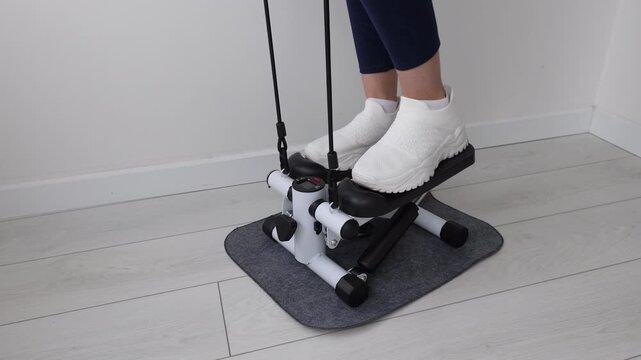 Close-Up of Woman's Feet in White Sneakers Using Mini Stepper Exercise Machine with Resistance Bands on Gray Mat at Home, Female Legs in Blue Leggings Working Out on Compact Stair Stepper Indoors