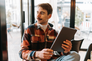 A stylish young man holding a tablet and looking thoughtfully out of a window while sitting in a cafe. © Dusko