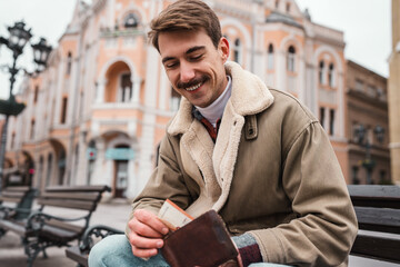 A smiling man with a mustache sitting on a bench in a city square, holding a banknote and a leather wallet while enjoying a casual moment. © Dusko