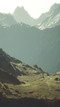 Mountains rise in the background with rugged features. The scene shows a mix of grass and rocky ground. Light casts shadows on the hills in a bright setting.