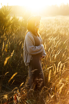 Stylish pregnant woman holding hands on belly and relaxing in sunlight in autumn park. Happy Mothers day! Beautiful mom hugging baby bump, and enjoying evening. Motherhood concept