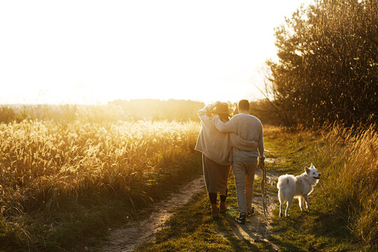 Beautiful pregnant couple walking with dog in sunset light in autumn park. Parenthood. Stylish happy young parents, mom and dad, enjoying evening and relaxing with pet