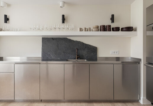 Modern minimalist kitchen with stainless steel cabinetry, open shelf displaying glassware and ceramic bowls.