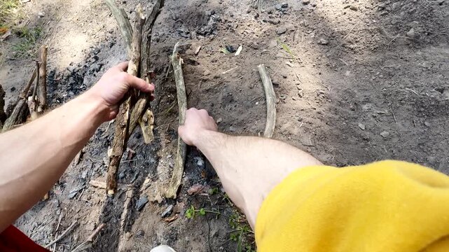 POV of a man in a yellow hoodie and red pants building a campfire frame from dry sticks on the ground. Primitive survival skills and bushcraft concept in the wild