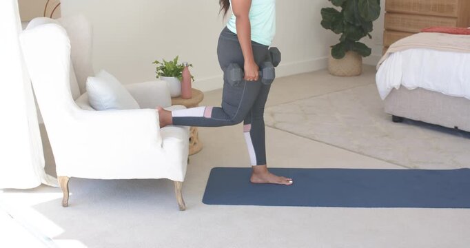 Woman in gymwear holding dumbbells foot on chair doing Bulgarian split squats on mat toning legs