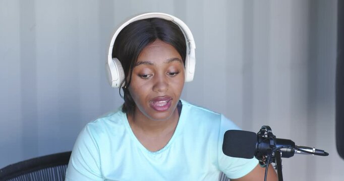 African American woman wearing headset reading notes speaking into mic recording podcast in studio