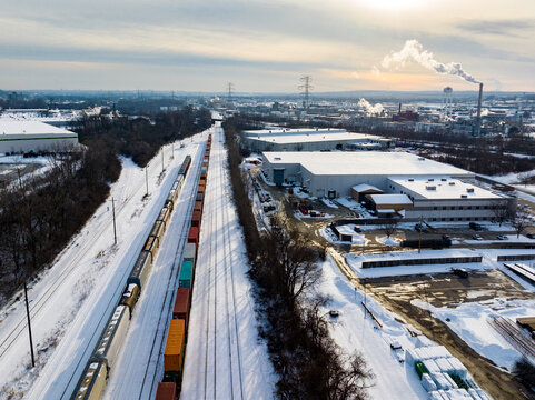 Aerial view of a freight train on snow-covered tracks passing through an industrial area with warehouses and rising steam under a winter sky Reading, Ohio, United States.
