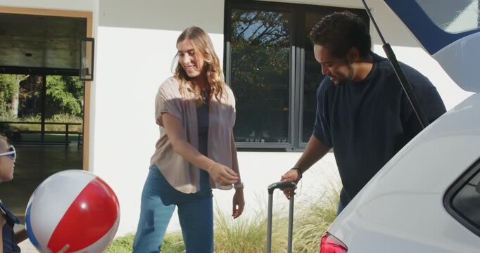 Family loading suitcase in driveway by car, parents handling luggage while child tossing beach ball