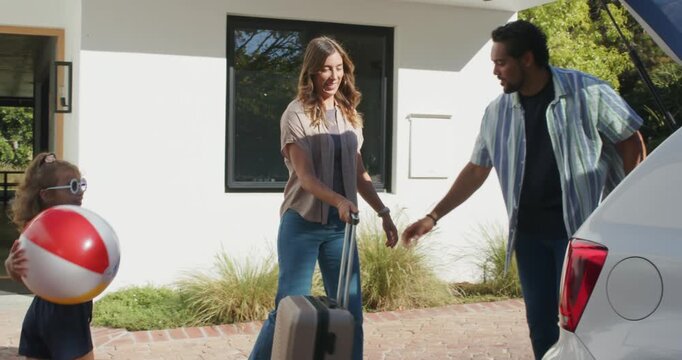 Family pulling suitcase, man loading trunk, woman kneeling tossing beach ball preparing to depart
