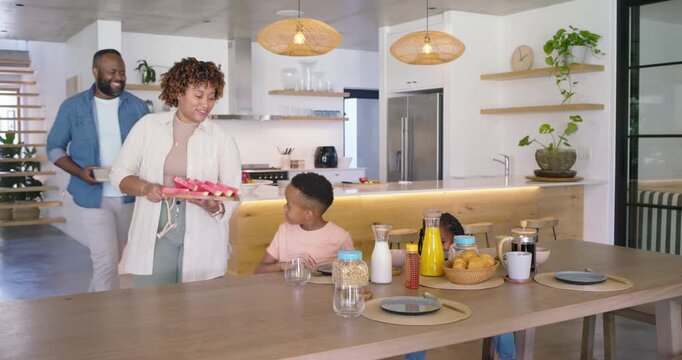 African American family preparing breakfast at table, mother pouring drink, offering watermelon