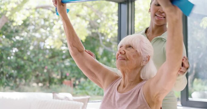 Caregiver placing hand on senior's upper arm, senior woman lifting blue band overhead for rehab