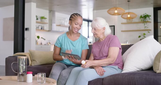 Caregiver pointing bottle and tapping tablet on sofa as senior woman reading label confirming dose