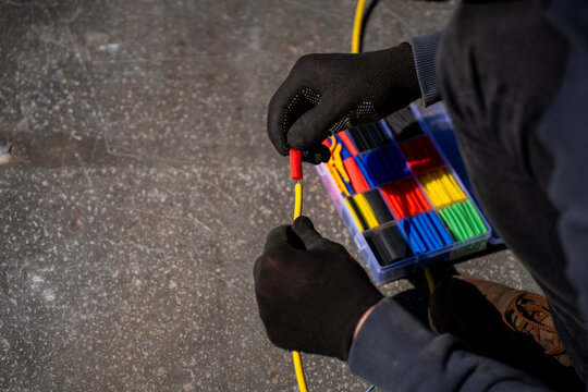 Gloved hands placing a red heat shrink tube over a yellow electrical cable beside a kit of colored shrink tubing outdoors.