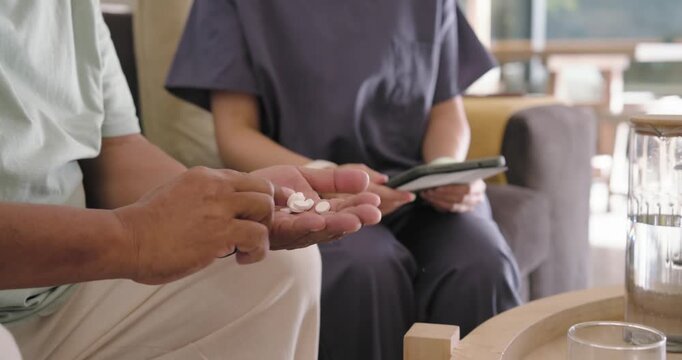 Asian caregiver in scrubs and man opening pill bottle on sofa counting, confirming dose on tablet