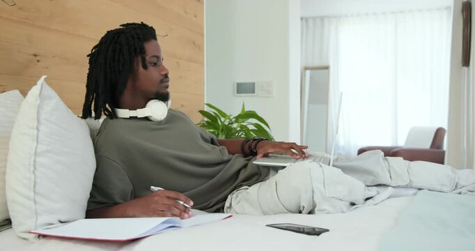 Man in twenties typing and writing in notebook on bed after seeing laptop screen, studying project