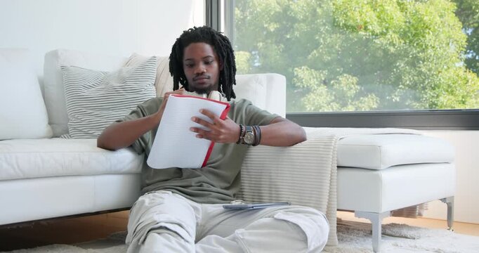 African American man studying on floor at home, writing red notebook, glancing at window, smiling