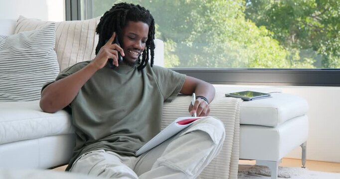 Adult African American man receiving call in living room, holding phone, taking notes in notepad