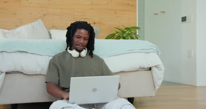 African man responding to smartphone ping, sitting on bedroom rug typing on laptop studying