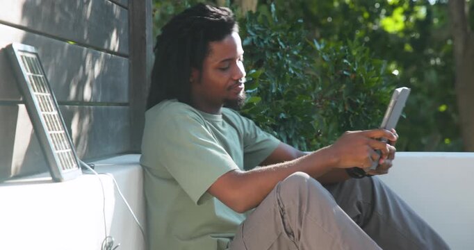 African man sitting on porch bench holding tablet, shifting grip, plugging solar cable to charge