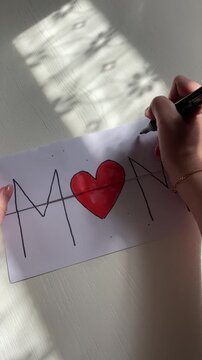 Vertical top-down video of a greeting card with &ldquo;M❤️M&rdquo; (heart replacing the letter O), random red marker dots being added around the design, Mother&rsquo;s Day card decorating process, soft natural lighting